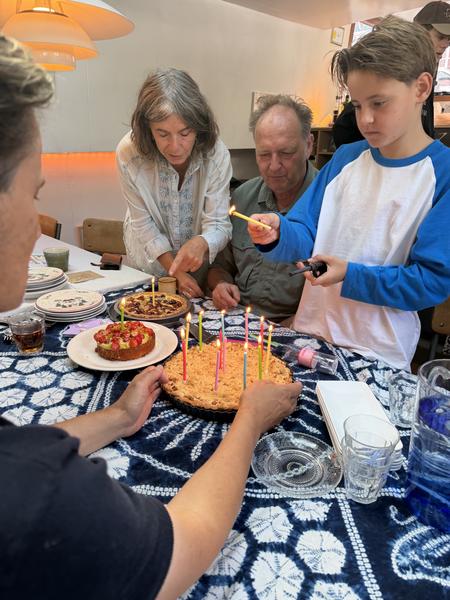 A family gathers around a table, lighting candles on birthday cakes in a warm indoor setting